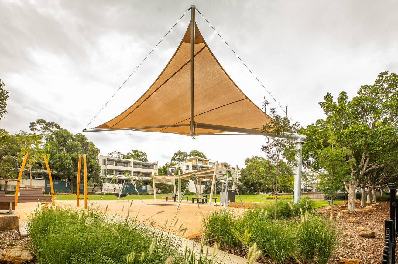 A large, single-post cantilever shade sail providing cover over a modern, landscaped community playground.