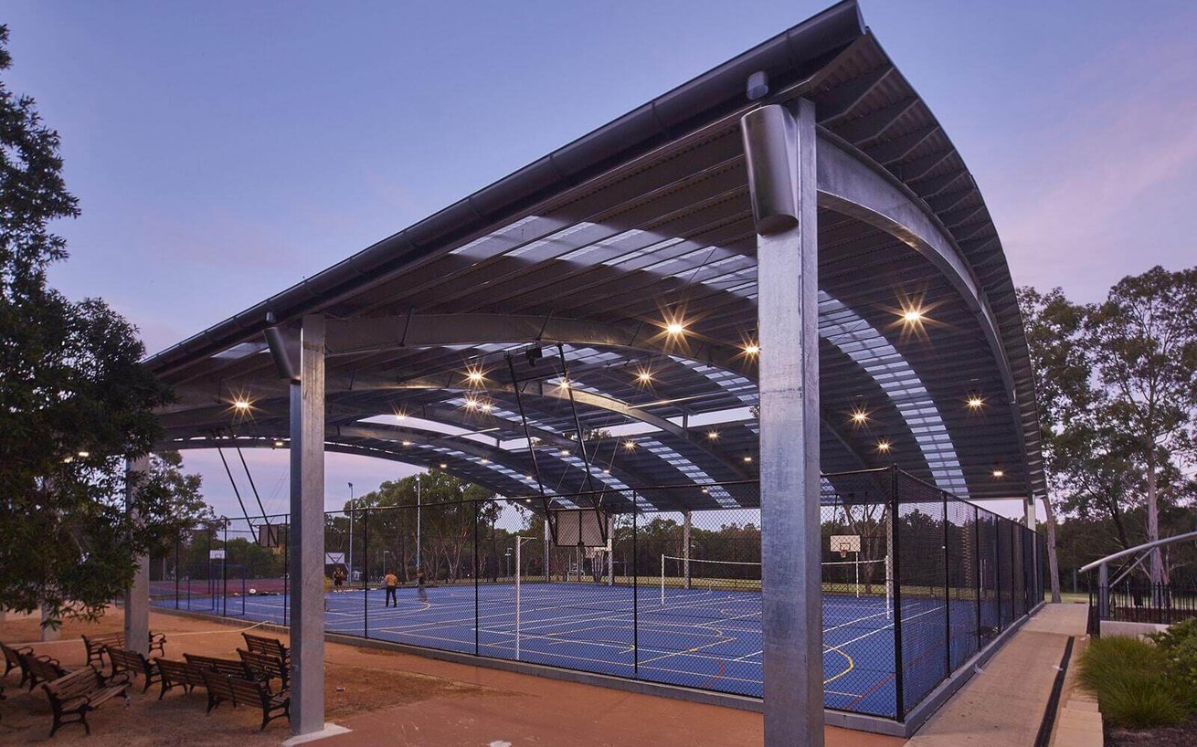 Large, illuminated COLA (Covered Outdoor Learning Area) with a steel-framed roof over a blue multi-sport court at dusk.