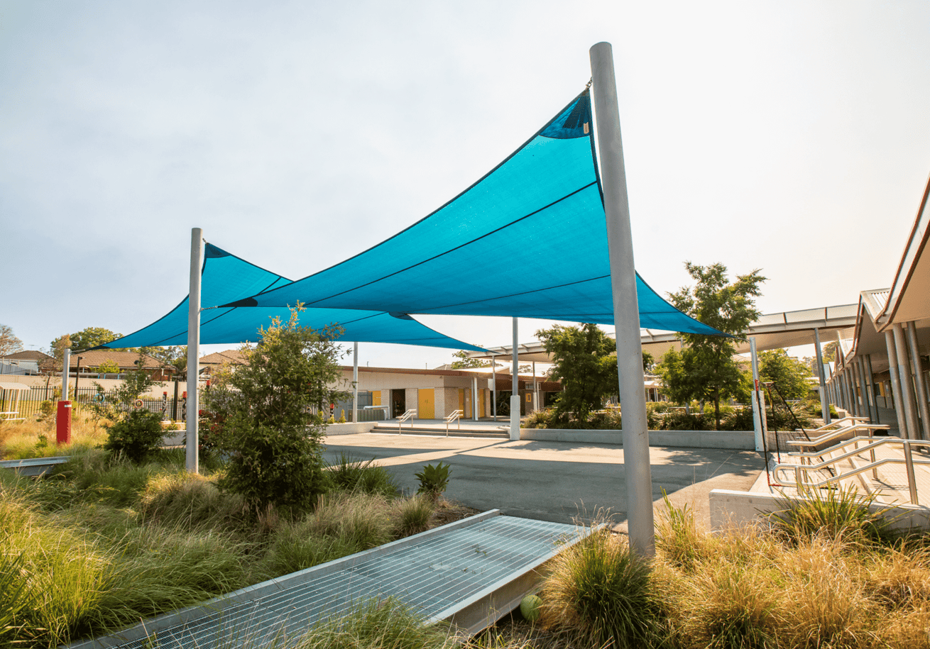 Bright blue architectural shade sails providing sun cover over a modern, landscaped school courtyard with native grasses.