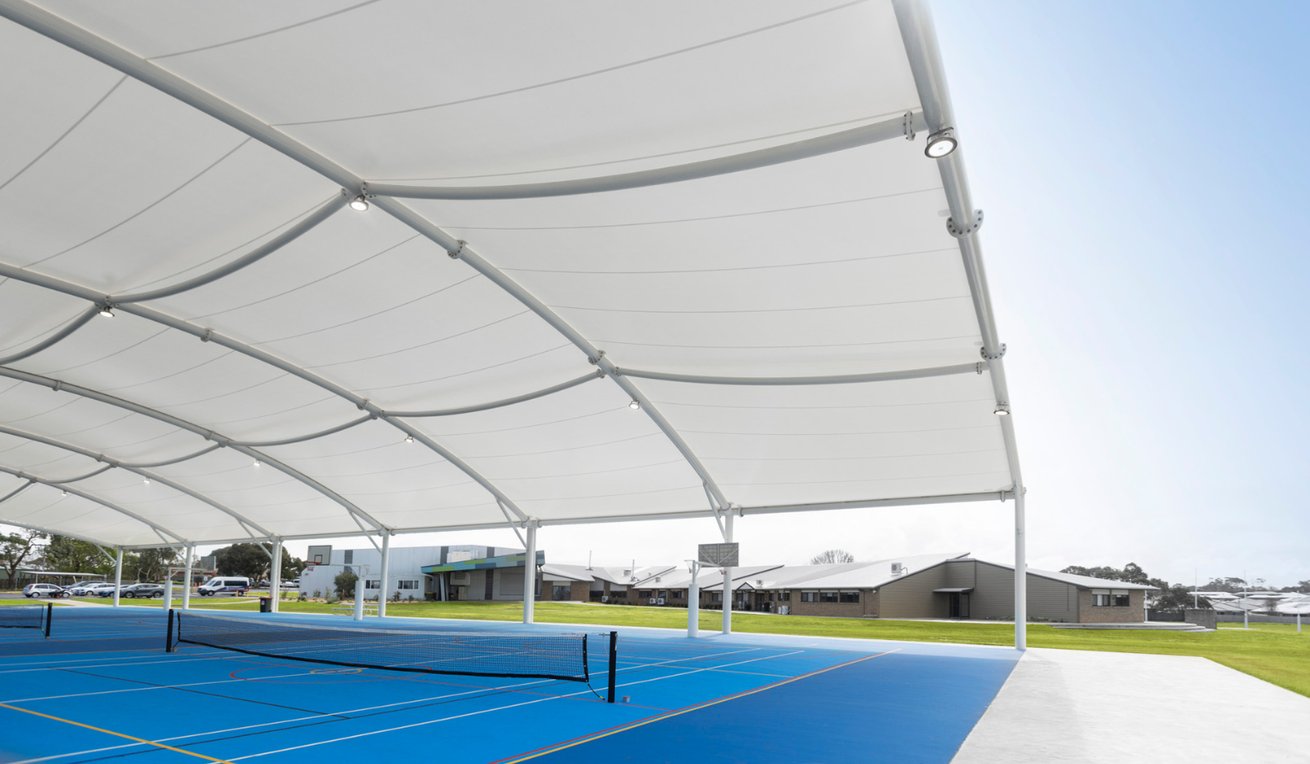 Underside view of a Greenline curved fabric shade structure with integrated lighting over a blue multi-use court at King's College, with tennis nets visible and school buildings in the background.