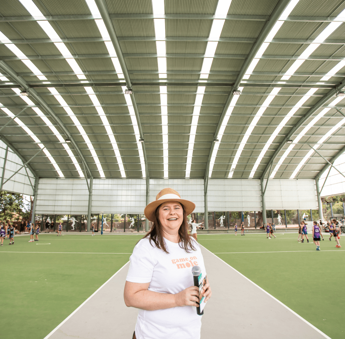A smiling woman standing under a metal roof structure with school kids playing netball in the background.